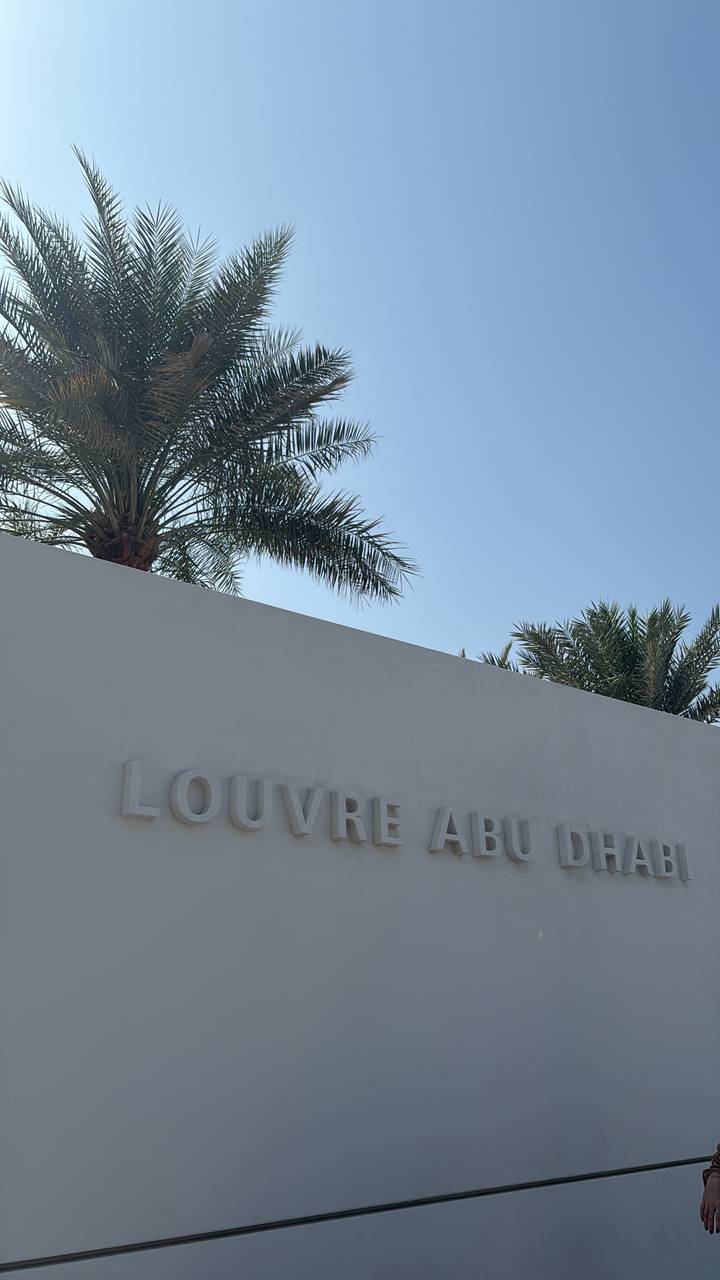 White exterior wall of the Louvre Abu Dhabi with raised lettering, framed by palm trees under a clear blue sky.