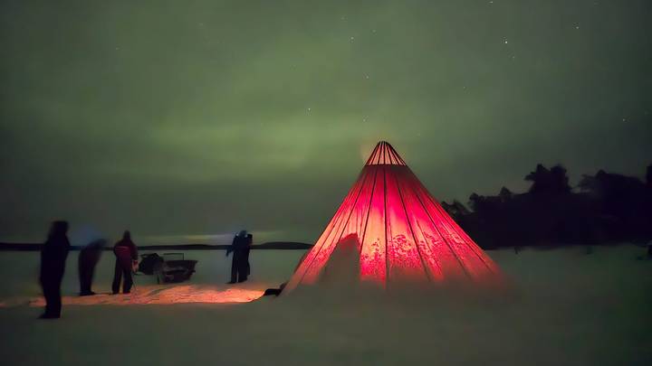Red canvas tepee glowing from within on snowy ground as silhouetted people observe the night sky.