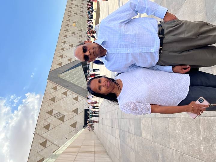 Mature couple posing outside the modern Grand Egyptian Museum with geometric facade under blue sky.