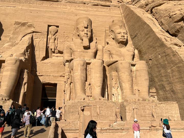 Massive seated statues of Ramses II at Abu Simbel with visitors gathered below in bright sunlight.