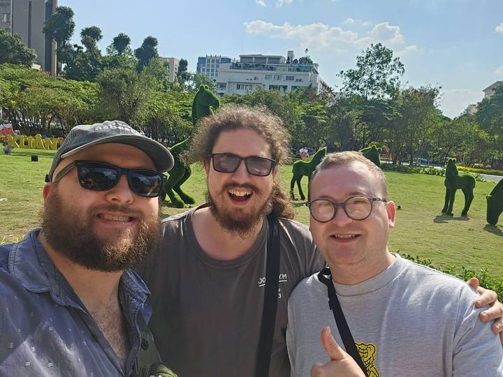 Selfie of three smiling travellers in a sunny urban park with topiary horse sculptures in the background.