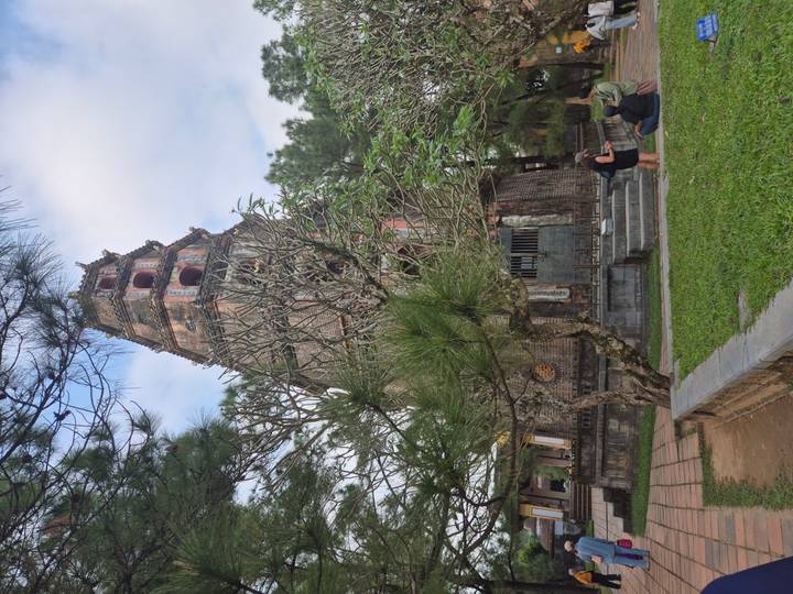 Tall ancient Thien Mu pagoda in Hue framed by pine trees with two visitors seated nearby.
