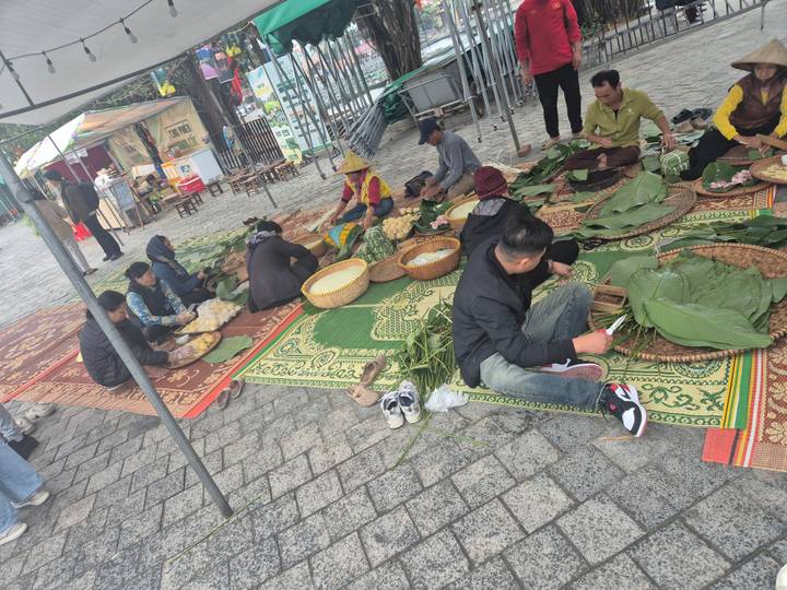 Local people sitting on mats preparing traditional food parcels with banana leaves in an outdoor market setting.