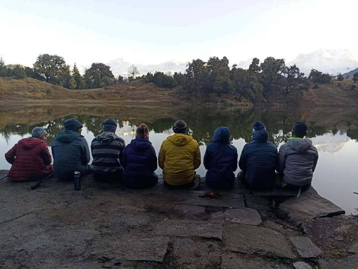 Trekkers in warm jackets sit side by side facing a still alpine lake with distant snowy peaks at dawn.