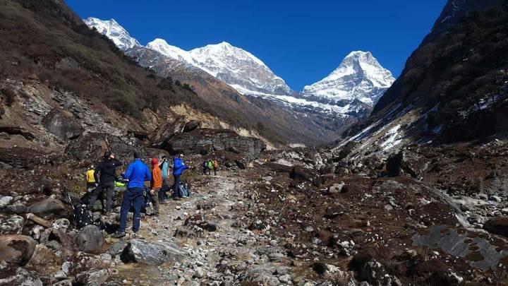 Hiking group traverses a rocky valley with towering snow-clad Annapurna peaks under a deep blue sky.