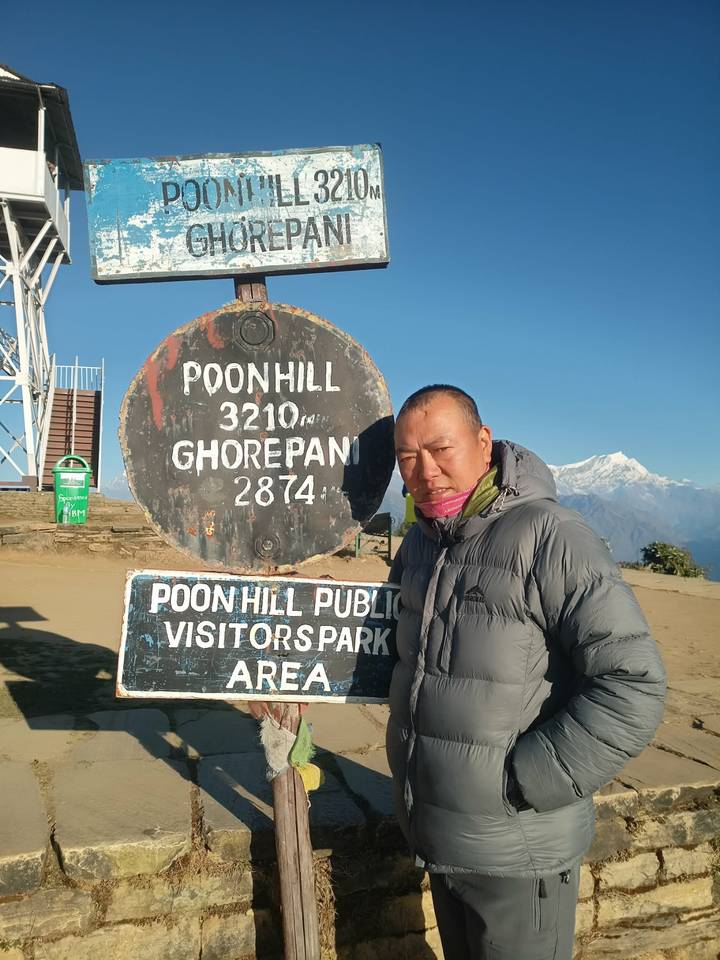 Trekker posing beside the weathered altitude signboard of Poon Hill with snowy Himalaya backdrop.