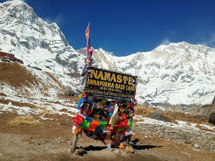 Bright prayer flags draped around the Namaste Annapurna Base Camp welcome sign against towering snow walls.