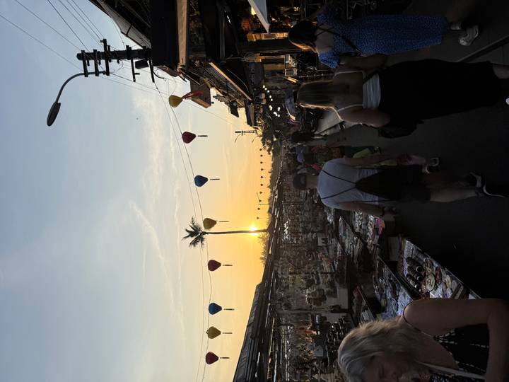 Bustling lantern-lit street market at sunset with shoppers walking beneath colorful hanging lanterns and a palm silhouette.