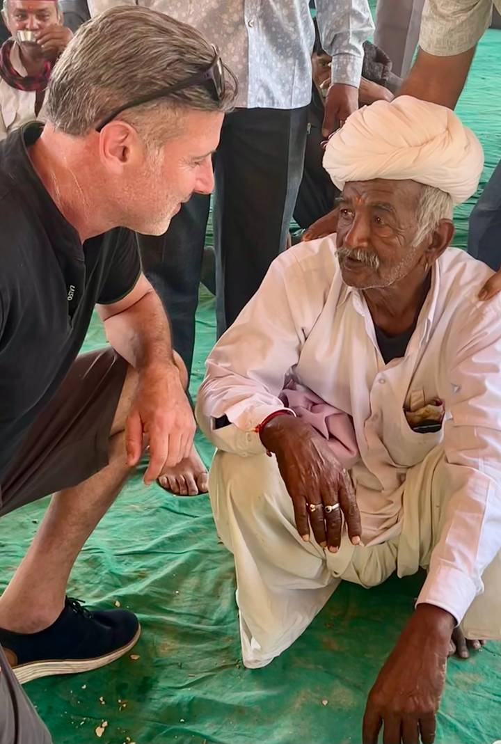 Close-up of a visitor crouching beside an older man in white attire during a communal gathering.