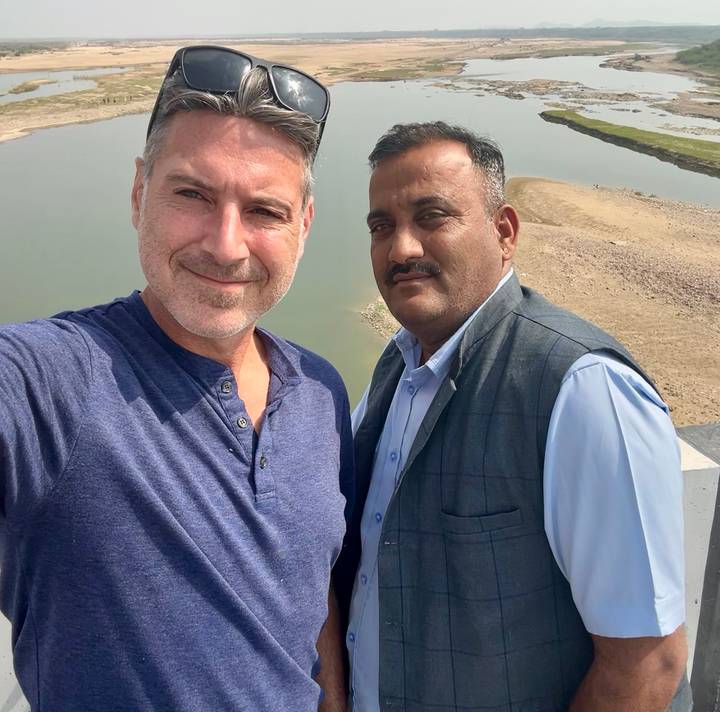 Selfie of two men standing on a bridge with a calm river and sandy bank behind them.