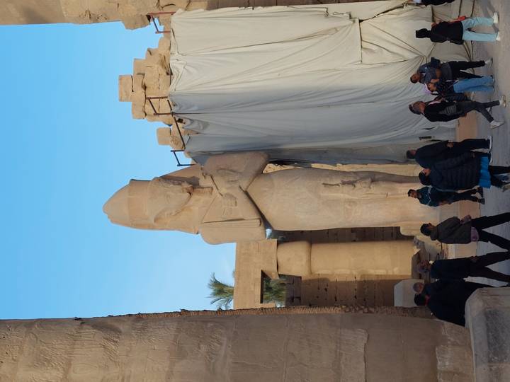Standing pharaonic colossus towers above visiting tourists inside an ancient temple courtyard.