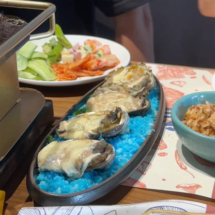 Cheesy baked oysters presented on vibrant blue salt beside a plate of vegetables and kimchi.
