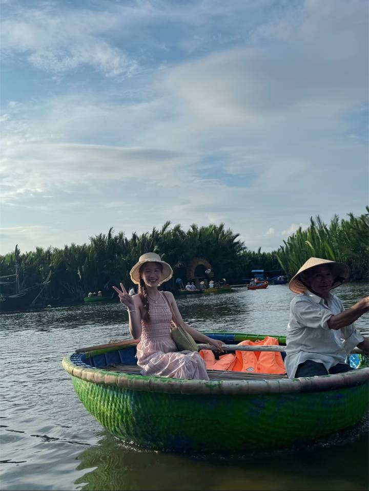 Traveller flashing a peace sign while sitting in a round basket boat rowed by a local wearing a conical hat on a nipa-palm lined river.