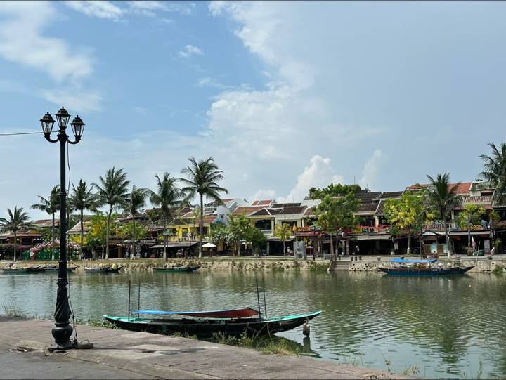 Charming riverside lined with palm trees and traditional shophouses reflected in calm water under a blue sky.
