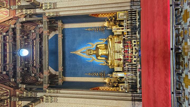 Gold Buddha statue in an elaborately decorated temple sanctuary with vivid blue backdrop and red carpet.