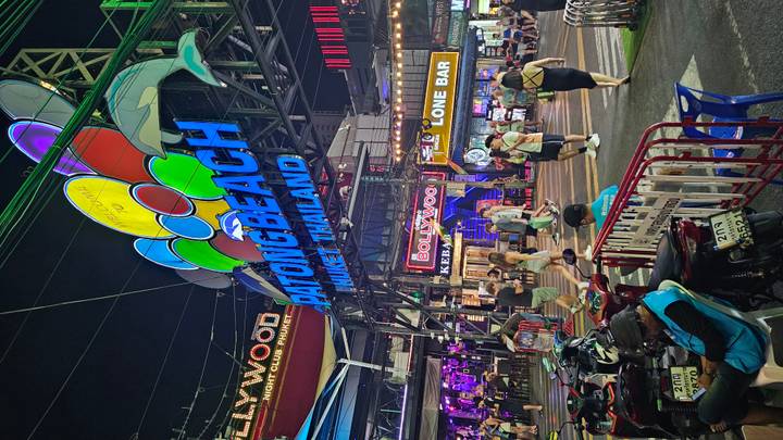 Neon night scene of Patong Beach sign with crowds, motorbikes and bars lighting up the street.