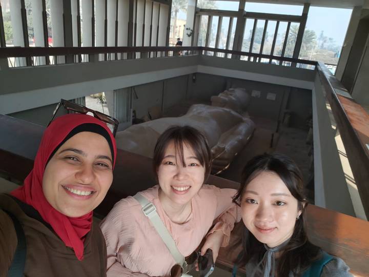 Smiling trio posing inside museum balcony overlooking colossal reclining statue of Ramses II.