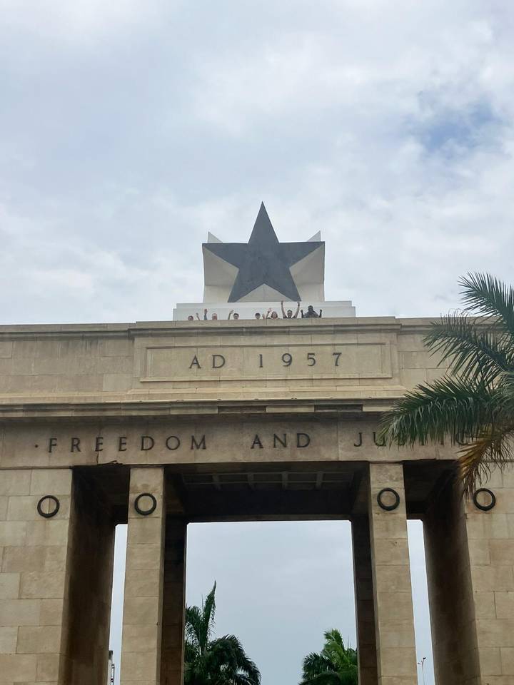 Black Star Monuments with tiny silhouetted visitors raising arms atop historic arch in Accra.