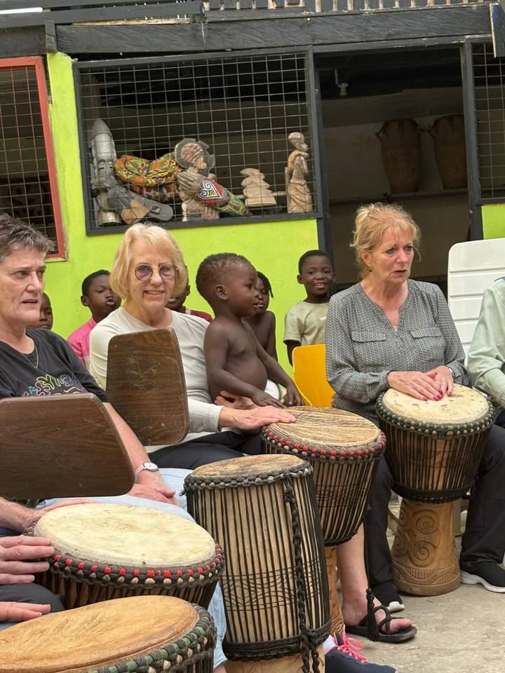 Visitors join local children in drumming session, holding traditional drums in bright courtyard.
