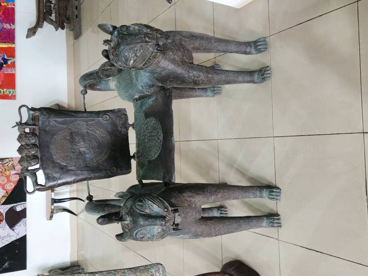 Bronze ceremonial chair flanked by two sculpted feline figures exhibited on a tiled floor inside a museum.