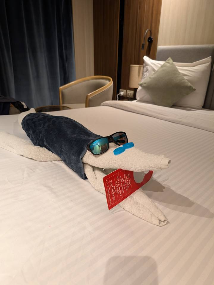 Playful towel animal shaped like a crocodile wearing sunglasses on a cruise ship bed.