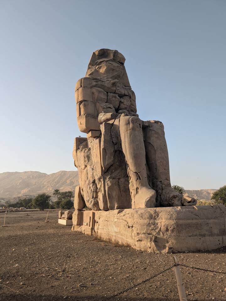 Massive seated Colossus of Memnon statue against arid mountains and blue sky.