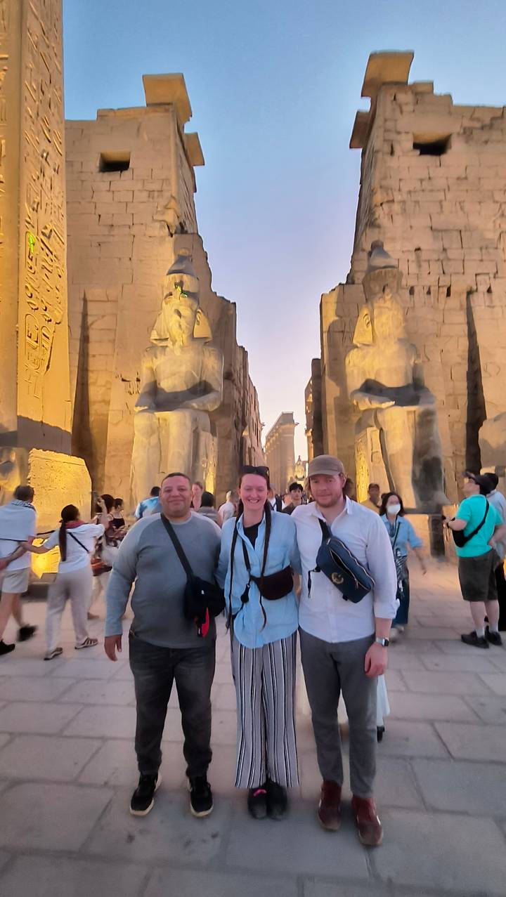 Travelers posing between colossal statues at the illuminated entrance of Luxor Temple at dusk.