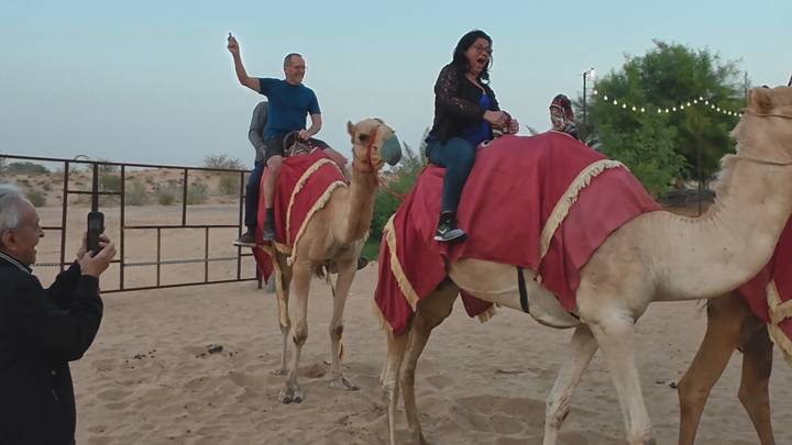 Travelers smile and pose while riding camels in the desert at dusk.