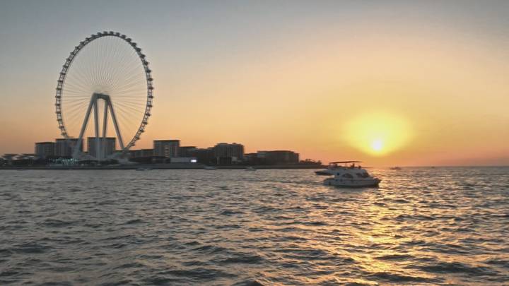 Ain Dubai ferris wheel silhouettes against a golden sunset as boats cruise the bay.
