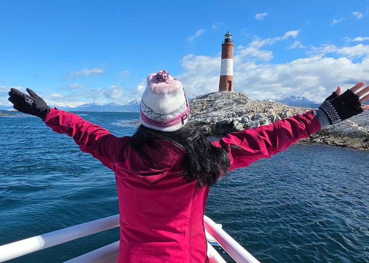 Traveler standing on a boat with arms spread wide facing the Les Eclaireurs Lighthouse in Ushuaia.