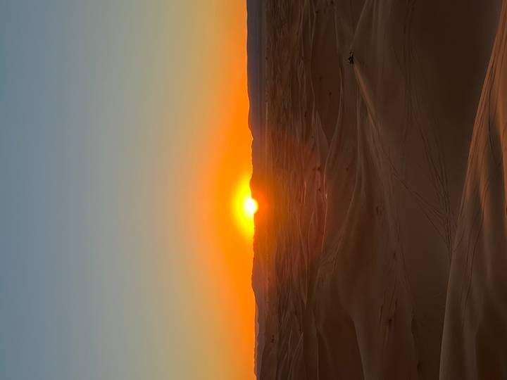 Brilliant orange sun sets over rolling Sahara dunes, casting long shadows.