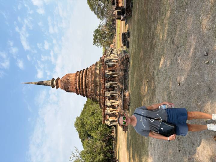 Traveler posing before an ancient brick stupa encircled by elephant statues in Sukhothai.