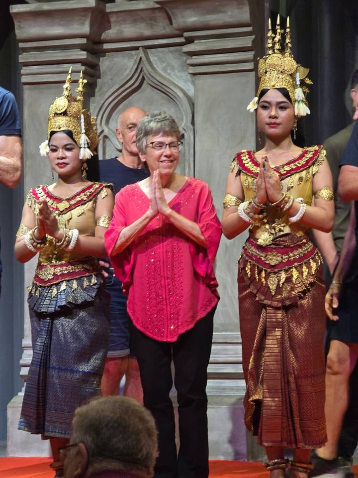 Traveler on stage posing with two traditional Apsara dancers in ornate Cambodian costumes