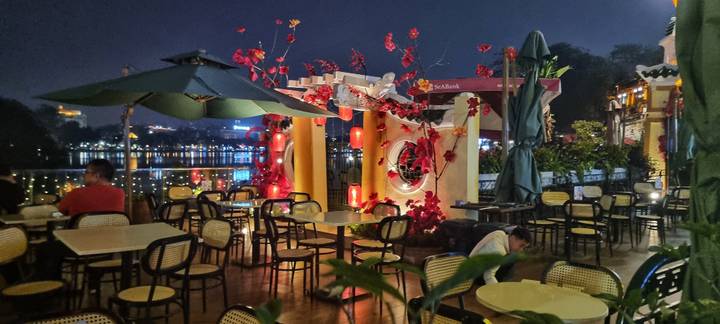 Riverside night cafe with red lanterns and empty tables overlooking city lights on the water.