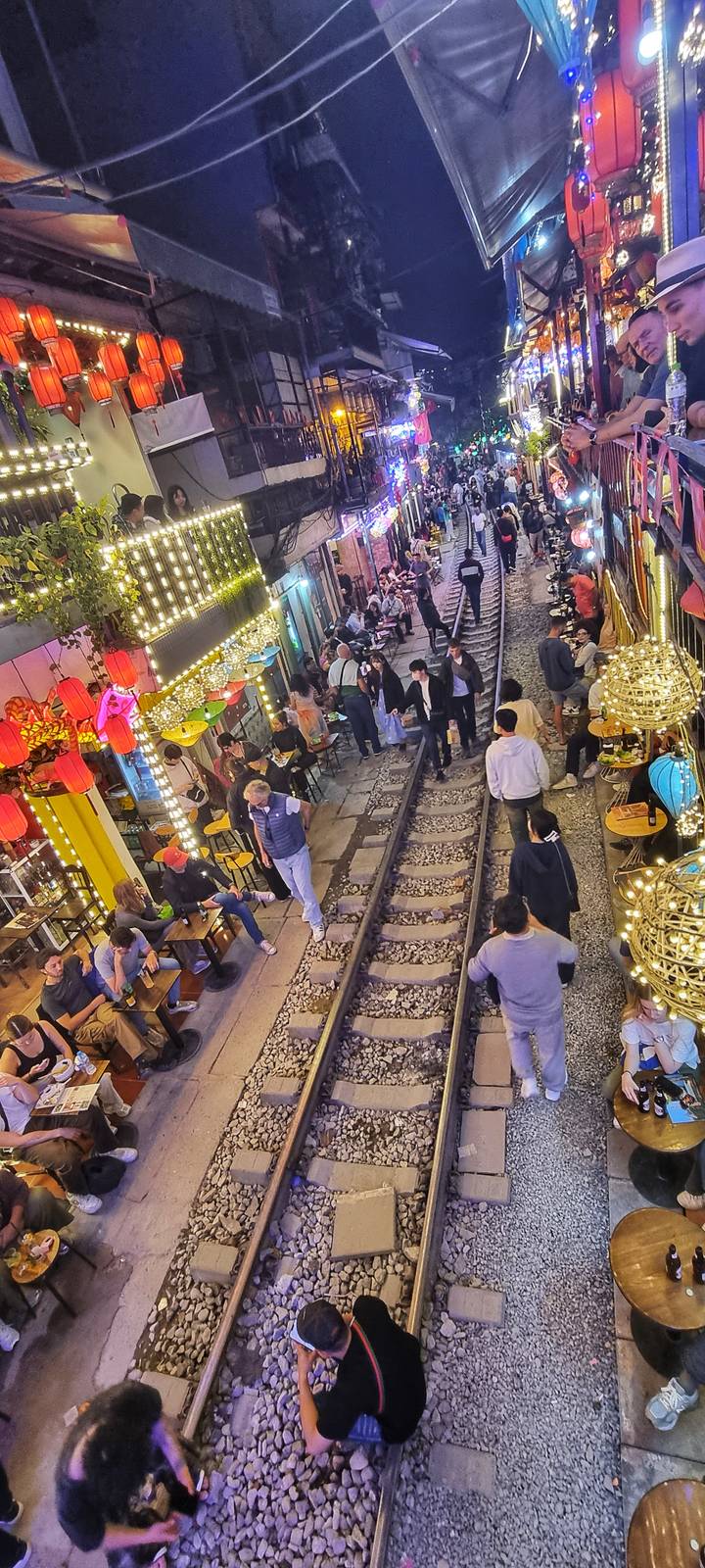 Bustling Hanoi Train Street lined with lit cafes and colorful lanterns as visitors sit beside the railway tracks.