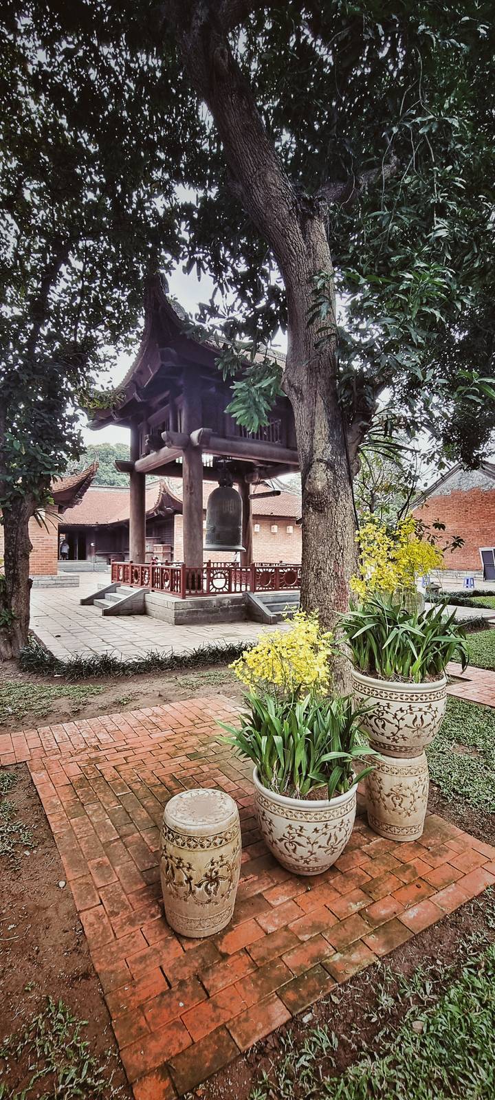 Large bronze bell hanging in a wooden pavilion inside a peaceful courtyard with yellow orchids.
