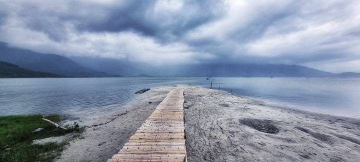 Narrow bamboo boardwalk extending into a tranquil bay under moody grey skies and distant mountains.