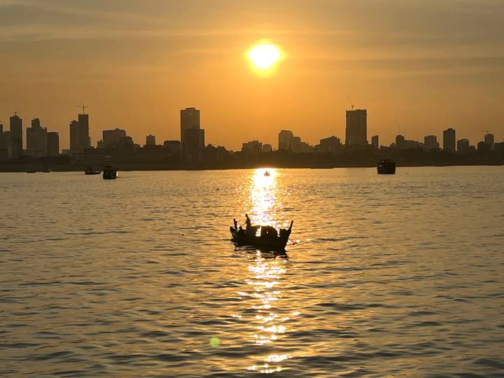 Silhouetted boaters raising hands on a river at golden sunset with city skyline in the distance