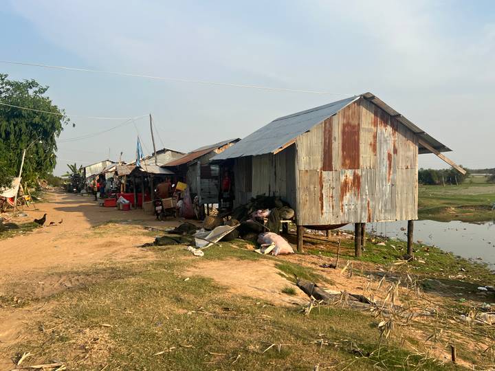Rustic corrugated-iron stilt houses line a dusty rural track beside a small pond and open fields