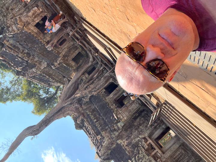 Close-up selfie with enormous tree roots engulfing temple walls at Ta Prohm under bright sky