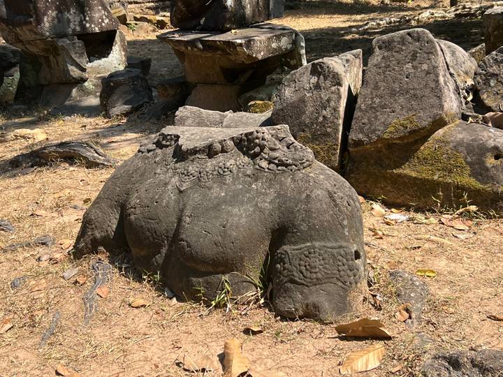 Weather-worn torso of a stone statue lying broken among boulders and dry grass in temple grounds