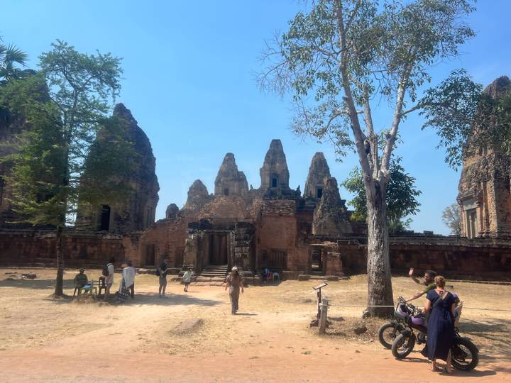 Ruined brick towers framed by tree and visitors exploring sunlit temple courtyard under blue sky