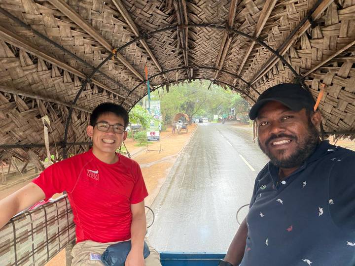 Smiling tourist and driver seated under a woven canopy tuk-tuk on a rainy road.