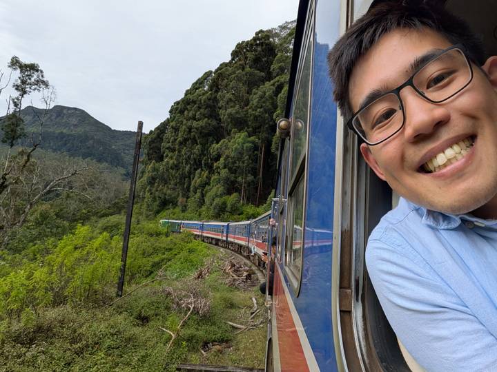 Smiling traveler leaning out of a blue train as it winds through lush green Sri Lankan mountains
