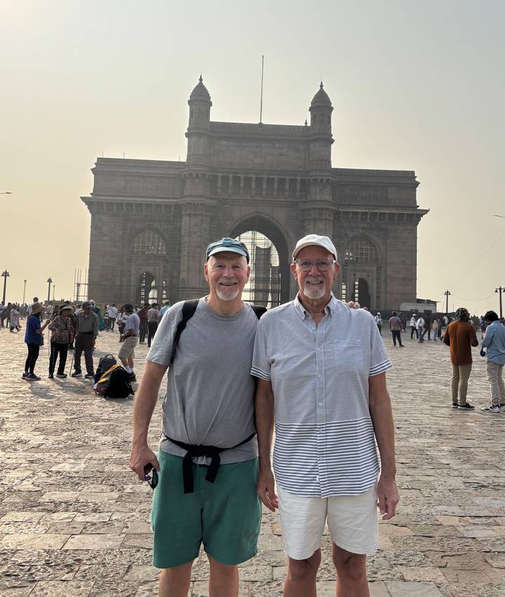 Travel duo stands before the imposing Gateway of India with crowds behind them