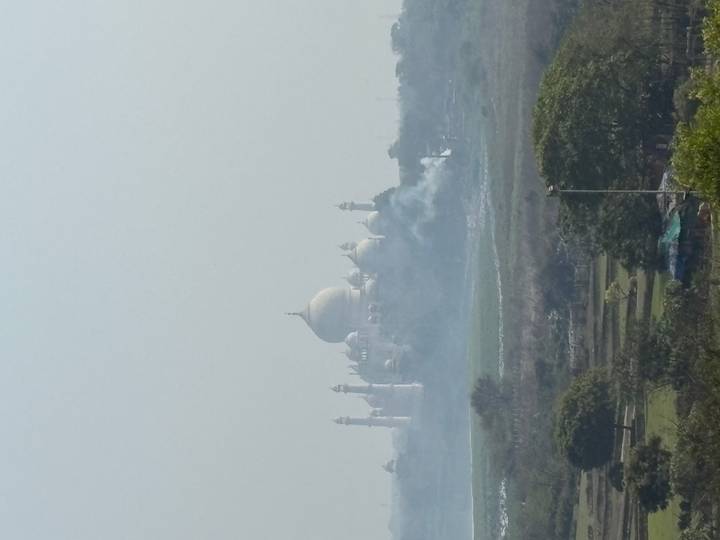 Distant, hazy view of the Taj Mahal beyond smoky fields under a pale sky.