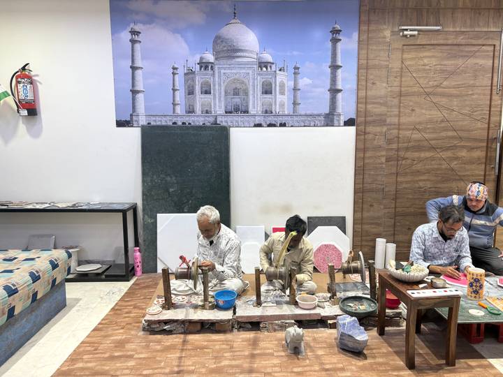 Artisans work on marble inlay pieces in a showroom with a Taj Mahal backdrop photo.
