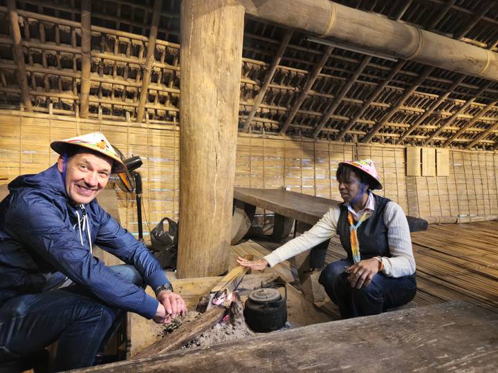 Travellers squat around a small hearth inside a bamboo stilt house during a cultural demonstration