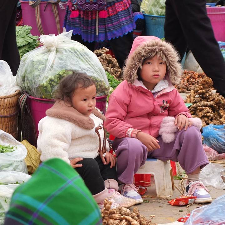 Two small children in warm clothes sit among produce baskets at a bustling Vietnamese market.