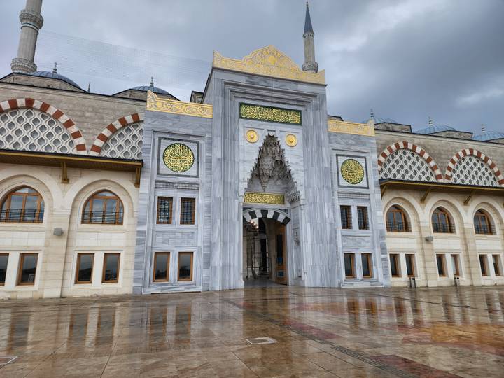Grand mosque entrance with ornate marble and gold details reflecting on wet courtyard.